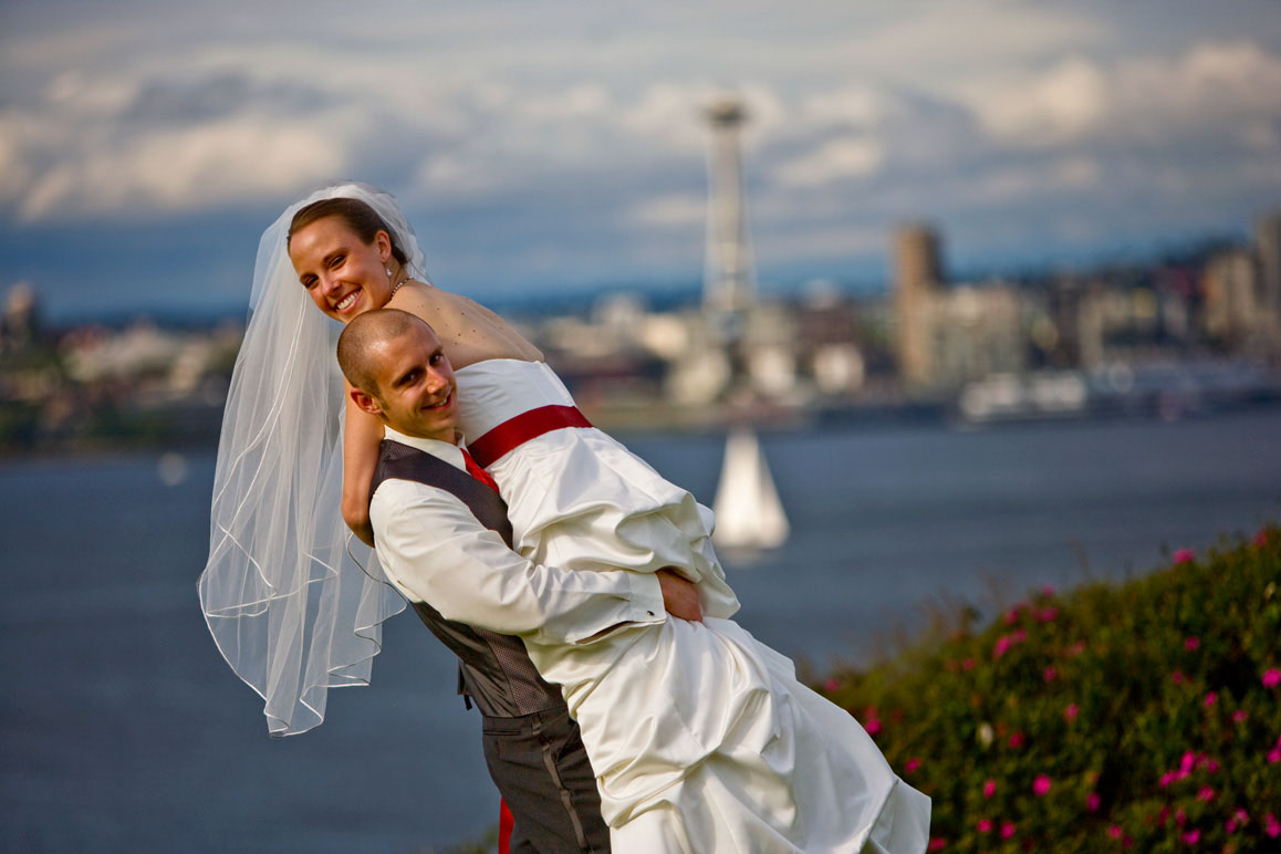 Wedding at Hamilton Overlook in West Seattle Groom carrying bride with Seattle and Space Needle in background in West Seattle